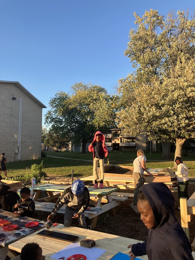 A child jumping for joy on a picnic table at Our Garden, with Art Club and Building Club around